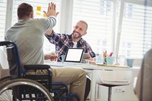 Dos hombres chocan las manos en un entorno laboral. El que está de frente sonríe y el que está de espaldas usa una silla de ruedas.