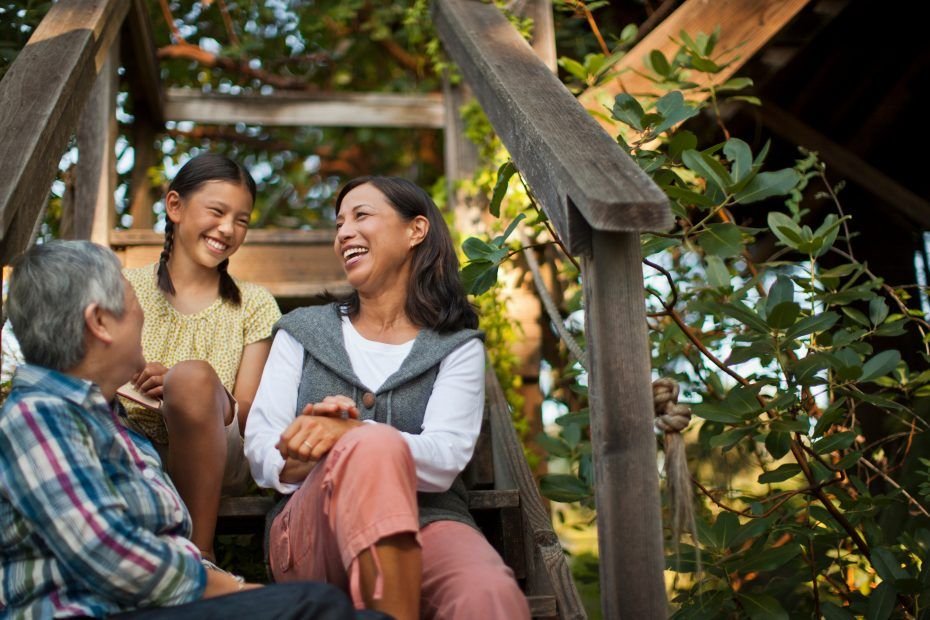 Dos mujeres adultas y una adolescente se sientan en unas escaleras de madera al aire libre. La mujer de mayor edad está de espaldas y mira a las otras dos. Las tres se ríen, con miradas de complicidad.