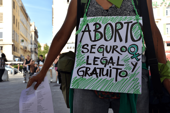 Torso de una persona que tiene un cartel colgado del cuello. El cartel dice «Aborto seguro, legal y gratuito».