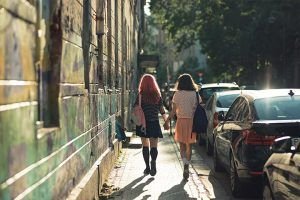 Dos mujeres de espalda, que caminan tomadas de la mano por una acera angosta. De un lado, están las fachadas de los edificios y del otro lado hay autos estacionados.