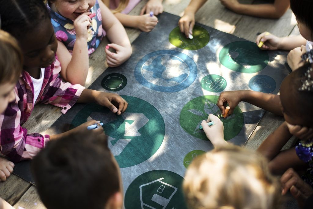 Un grupo de niñas y niños se reúnen alrededor de una mesa y escriben con tiza en una pancarta con símbolos ambientales.