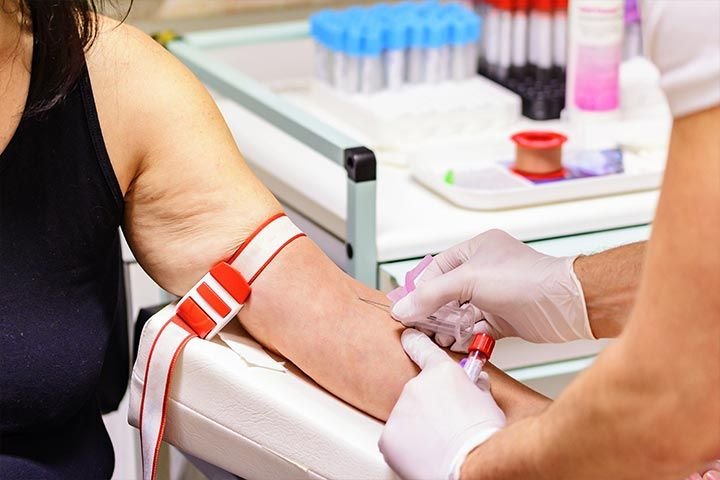 Foto del brazo extendido de una persona. Otra persona con guantes le está insertando una aguja en la parte interna del brazo. En el fondo, una mesa de laboratorio con varios tubos que contienen muestras de sangre.