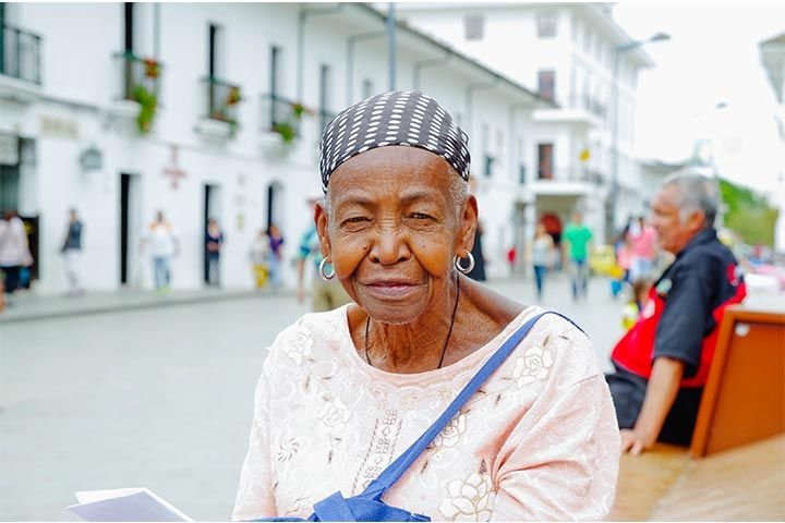 Adulta mayor afrolatina, con aretes y pañuelo a lunares en la cabeza, mira a la cámara. En el fondo difuso, se ve una calle y edificios blancos.