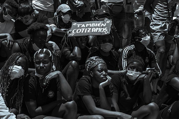 Grupo de jóvenes afrolatinos sentadas y sentados sobre gradas. Algunas personas usan mascarillas y hay un cartel que dice «también cotizamos».