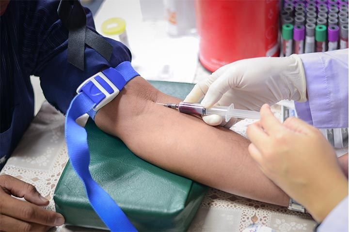 Foto del brazo extendido de una persona. Otra persona con guantes le está insertando una aguja en la parte interna del brazo. Al fondo, varios frascos de laboratorio con muestras de sangre.