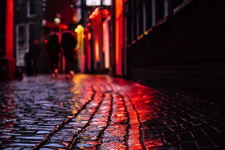 Primer plano de los adoquines de una calle en la ciudad. Es de noche, el suelo está mojado y se ve el reflejo de las luces rojas de unos carteles.