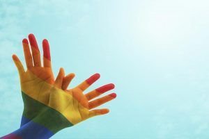 Dos manos entrelazadas por el dedo pulgar contra un cielo celeste. Las manos están pintadas con el color del arcoíris.