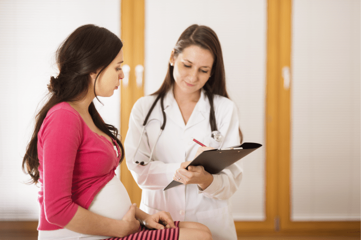 Mujer embarazada de perfil está sentada junto a una médica que lleva un estetoscopio en el cuello y sostiene una carpeta.