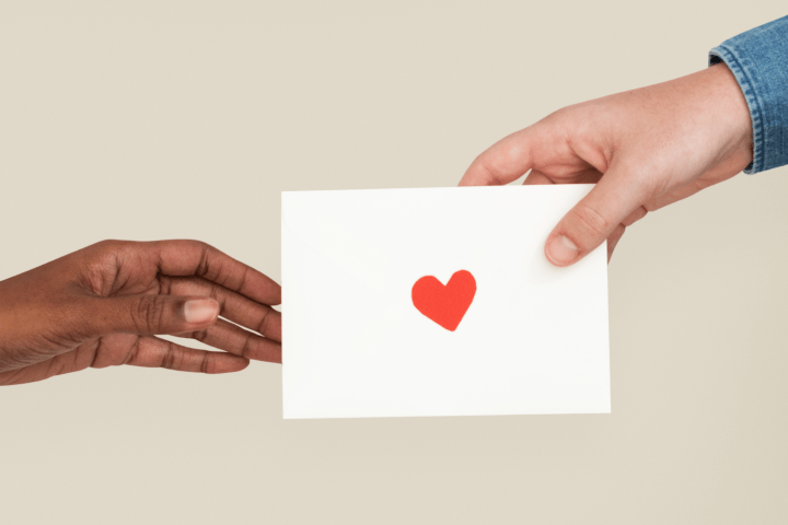 Imagen de la mano de una persona entregando un sobre a otra. El sobre tiene una pegatina de corazón.