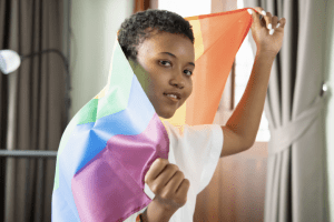 Plano medio de una joven que mira a cámara y sostiene una bandera del arcoíris sobre su espalda.